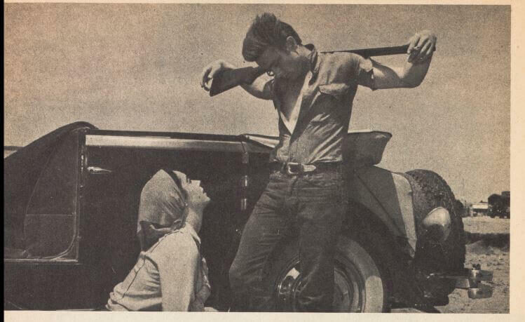 A man in a tank top flexes his muscles while a woman looks up at him, near an old car