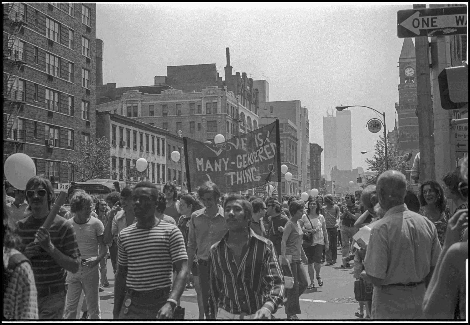 A crowd walking in a parade, holding a large banner that reads 'We Are Many, We Are Everything'. Balloon decorations and buildings are in the background