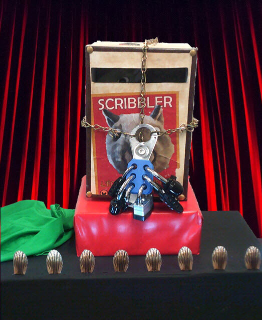 A mixed media display features a book titled "Scribbler" with an abstract sculpture in front, surrounded by small wooden pieces