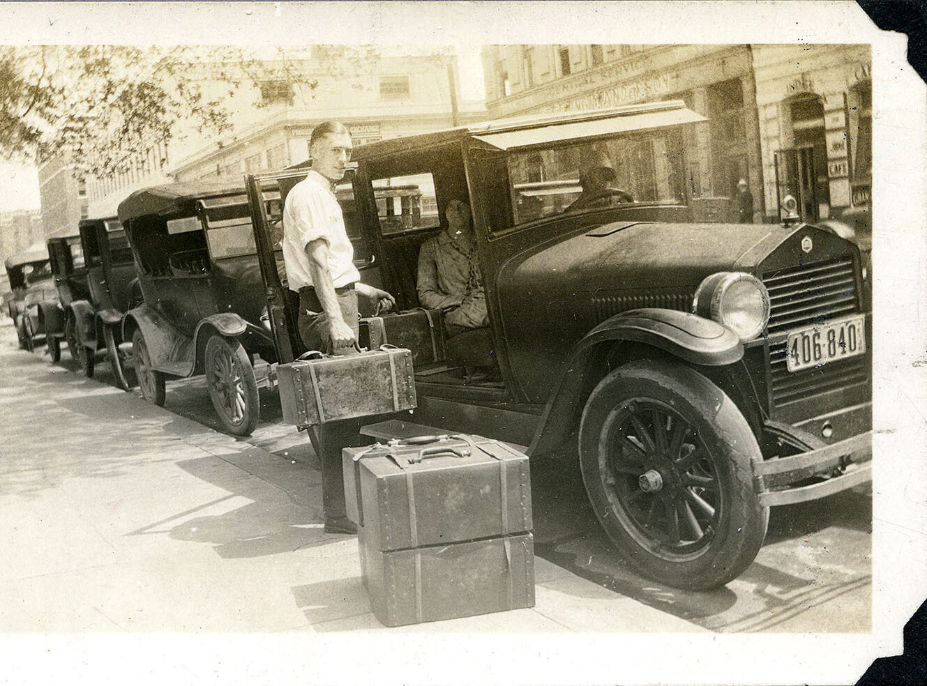 A vintage black and white photo of old cars parked on a street, with a man loading luggage.
