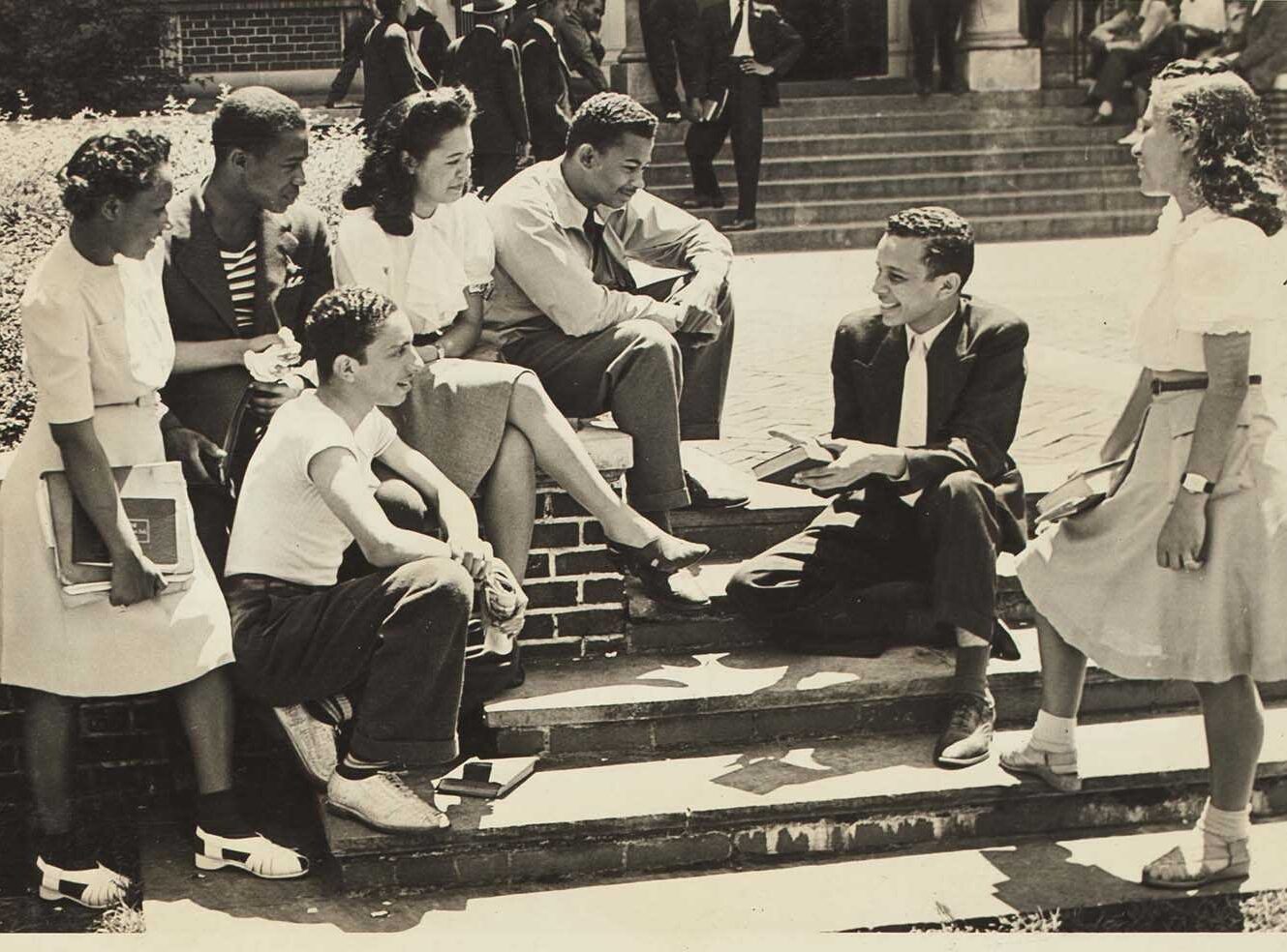 A group of six Black students sitting on steps, engaged in conversation and reading books.