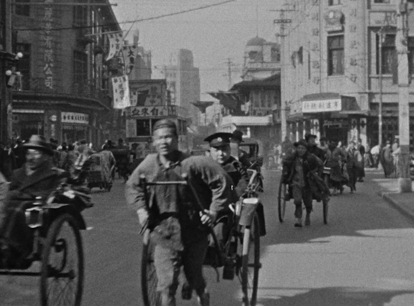 A grayscale busy street scene with rickshaws and pedestrians. Signs and buildings line the road.