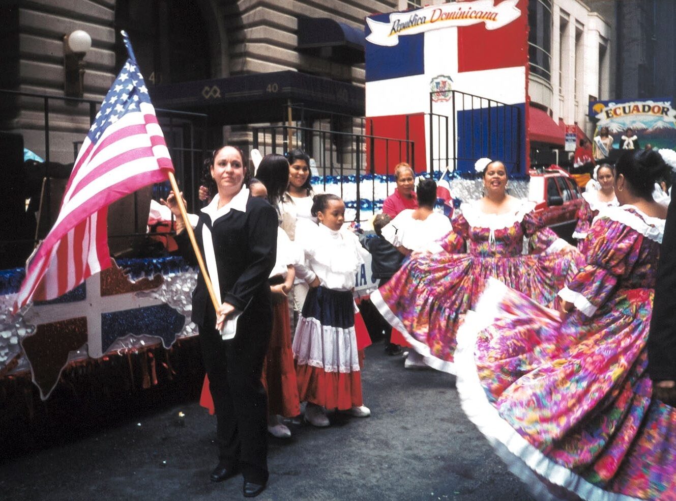 A colourful parade with women dancing and holding American, Dominican Republic and Ecuadorian flags.