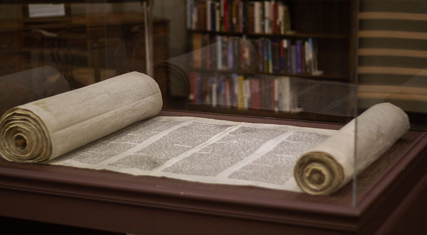 Two open scrolls on a display case with text visible, surrounded by books.