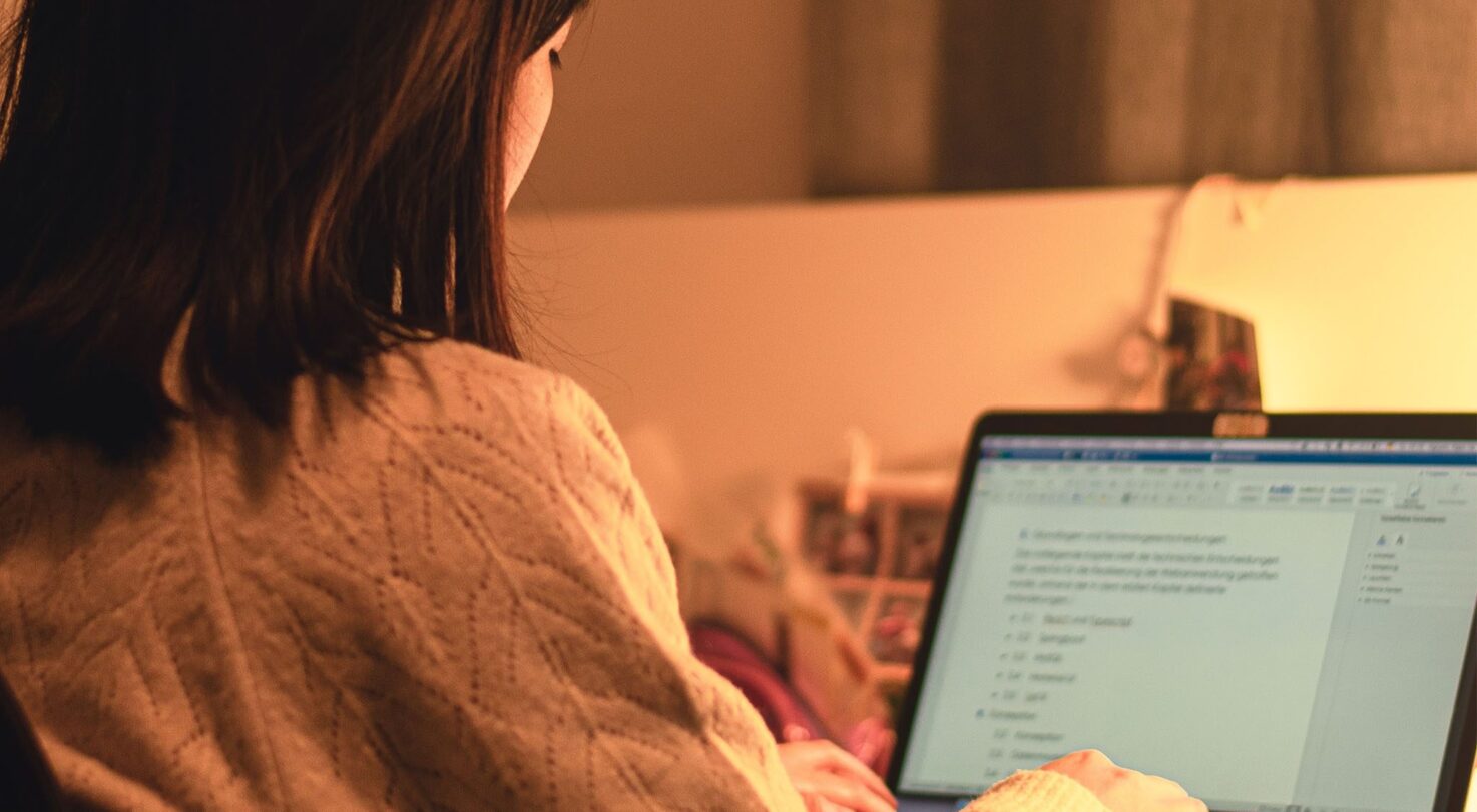 A person with brown hair sits at a desk, typing on a laptop with a document open.