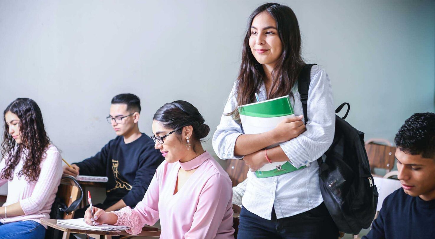 A classroom with students seated at desks. A girl stands holding green notebooks while smiling.