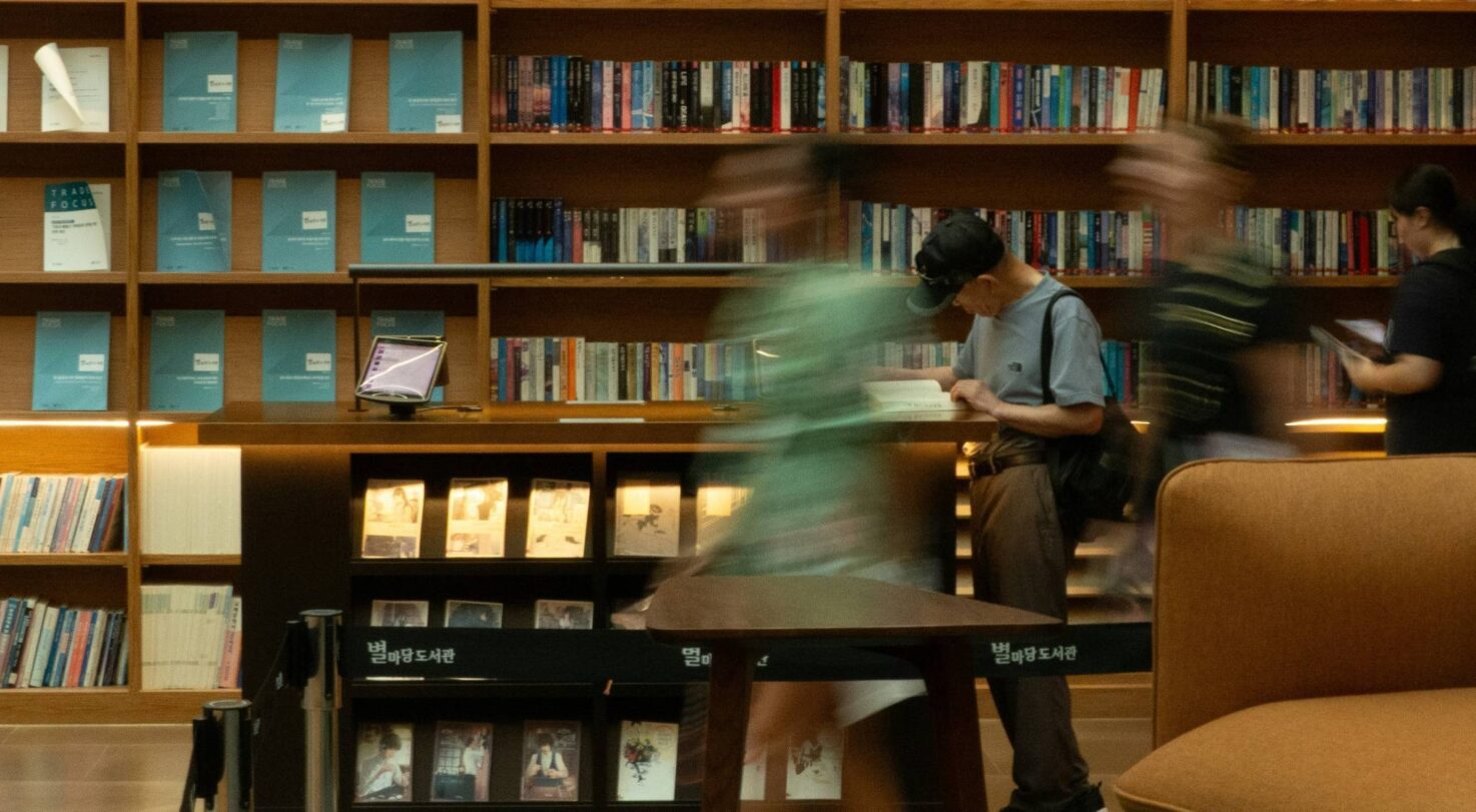 A busy library with a wooden bookshelf full of books and people browsing. A table in the foreground.
