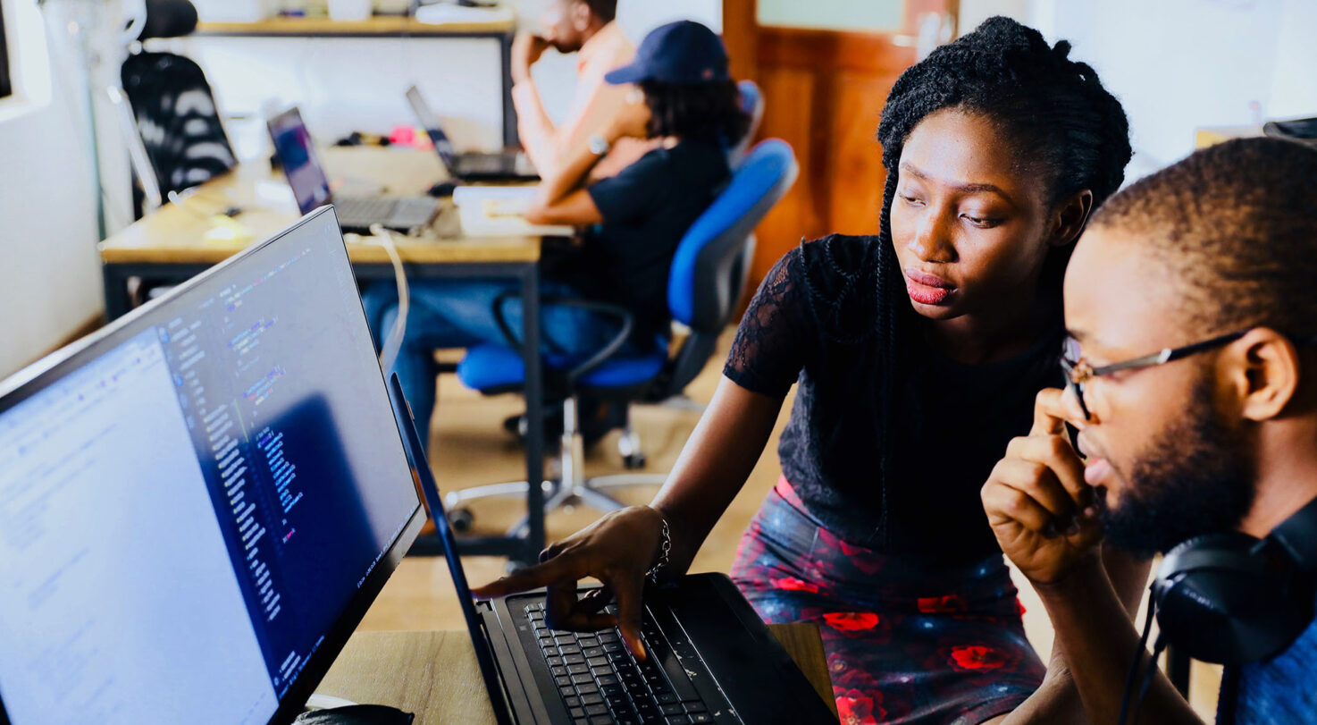 A woman points to a laptop screen while discussing with a man. Other people work on laptops in the background.