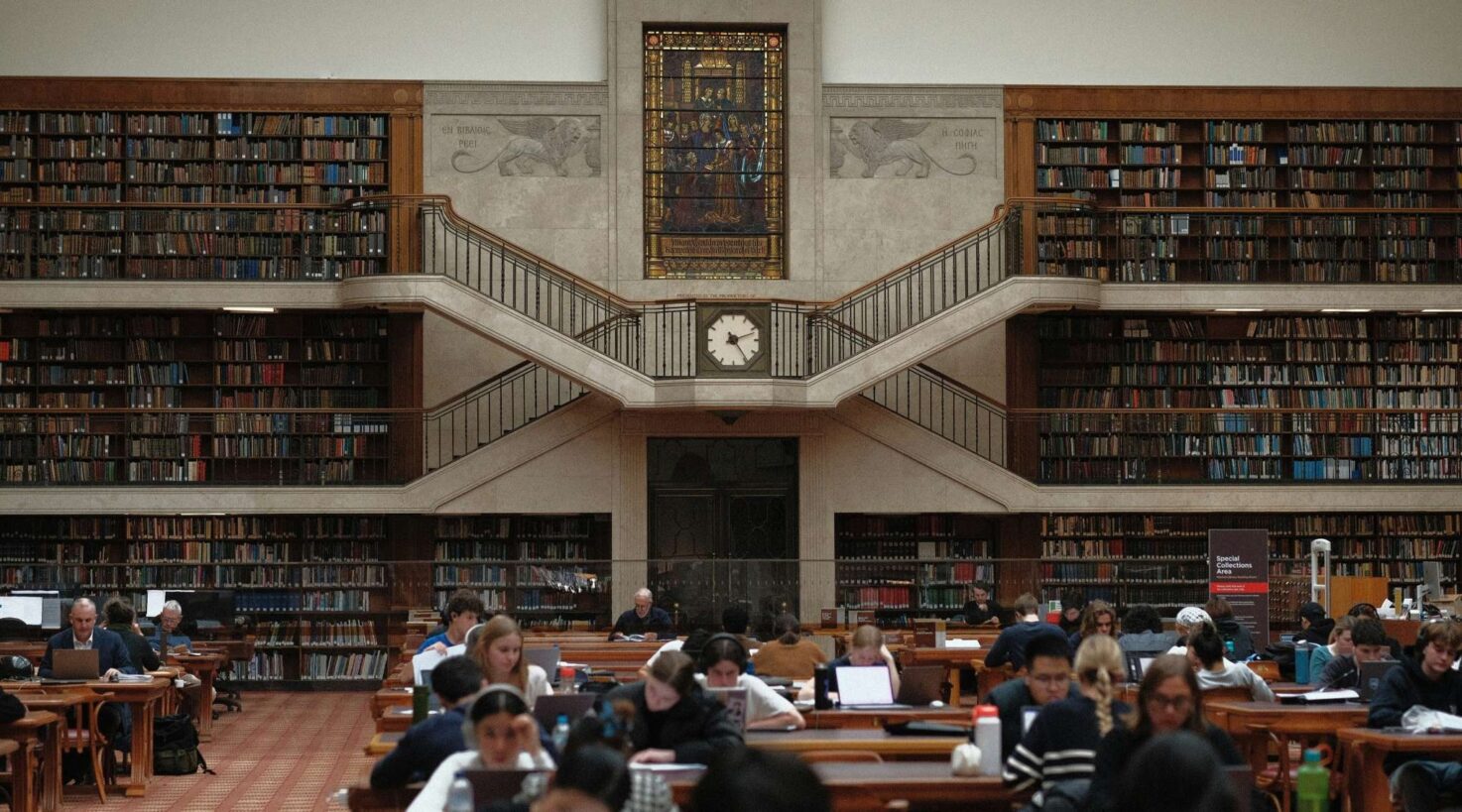 Inside a library with tall bookshelves and students studying at tables. A large clock and stained glass window are featured.