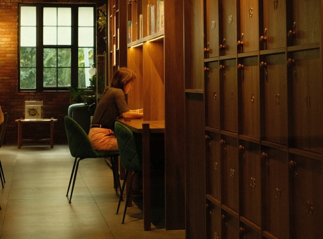 A woman sits at a wooden desk in a cosy, well-lit space with bookshelves and lockers.