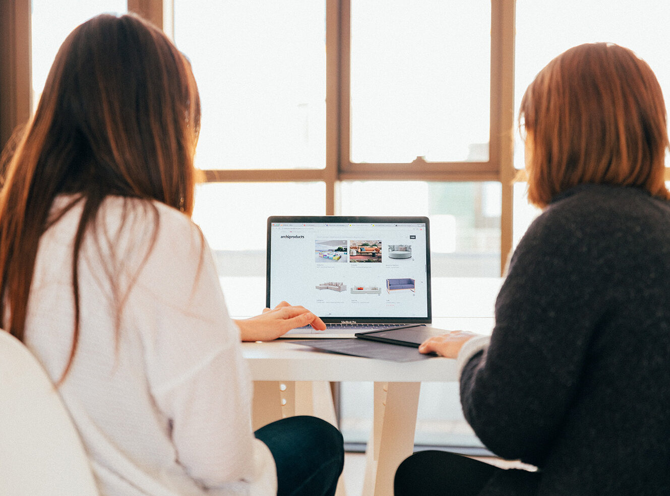 Two people sitting at a table, looking at a laptop showing furniture products.
