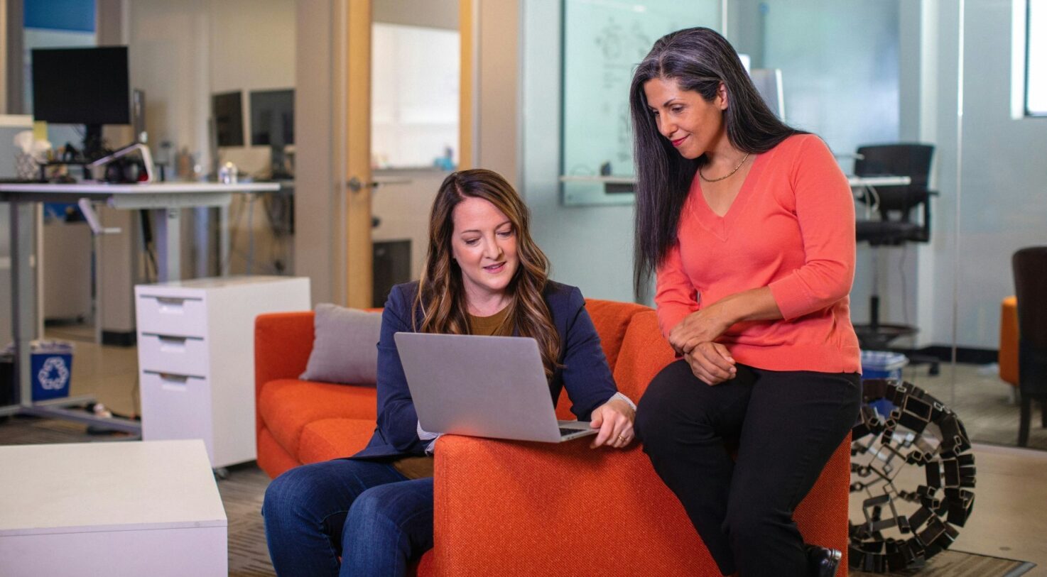 Two women in an office discussing over a laptop on an orange couch.
