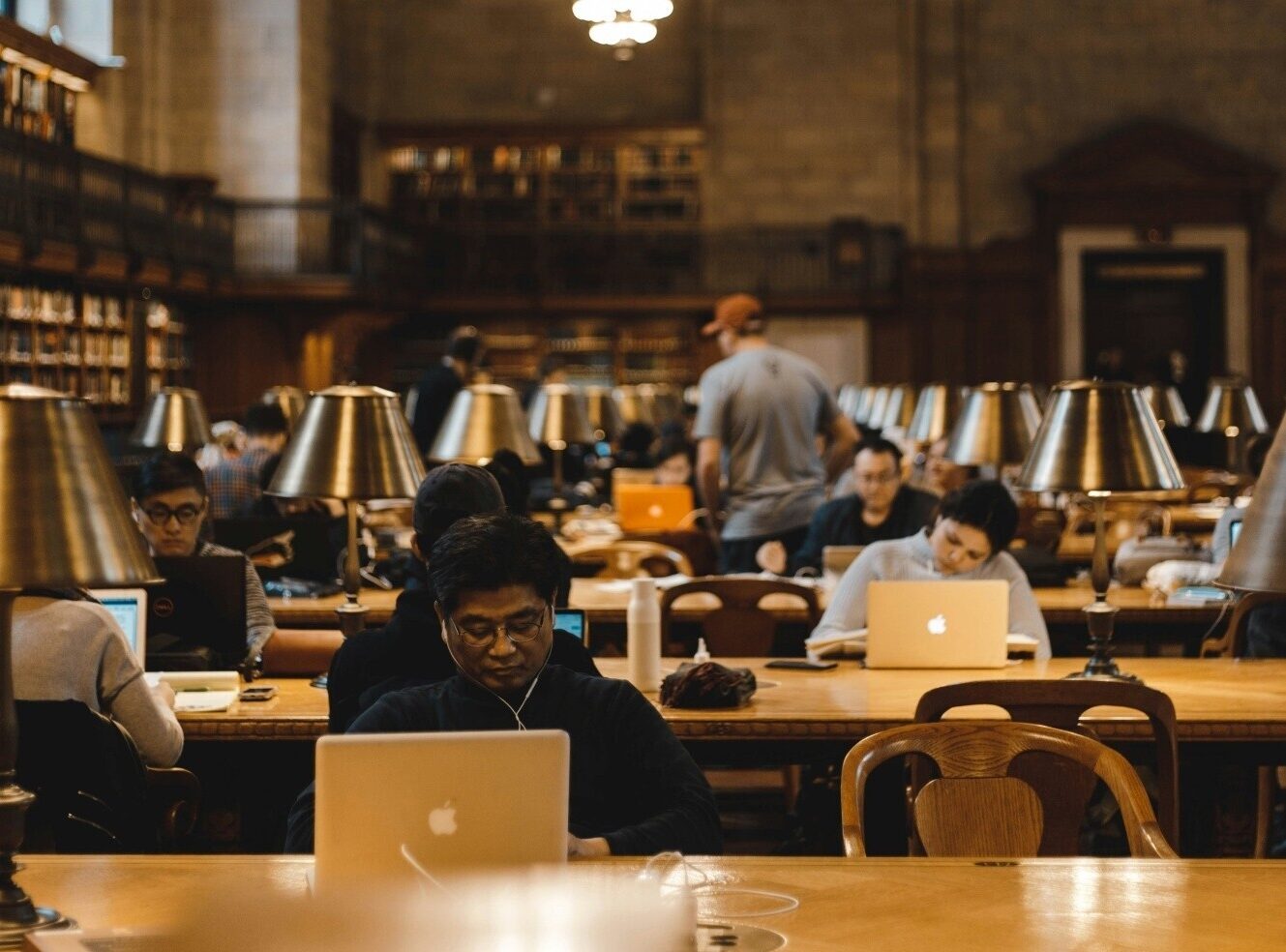 A busy library with people studying at wooden tables and using laptops. Desk lamps are lit.