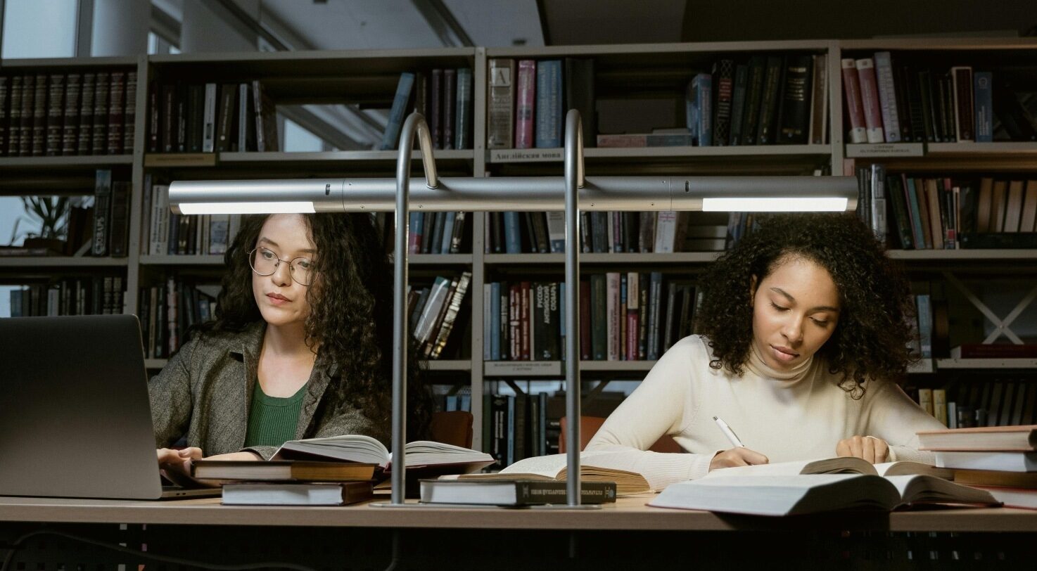 Two young women studying in a library, using laptops and books on a table.