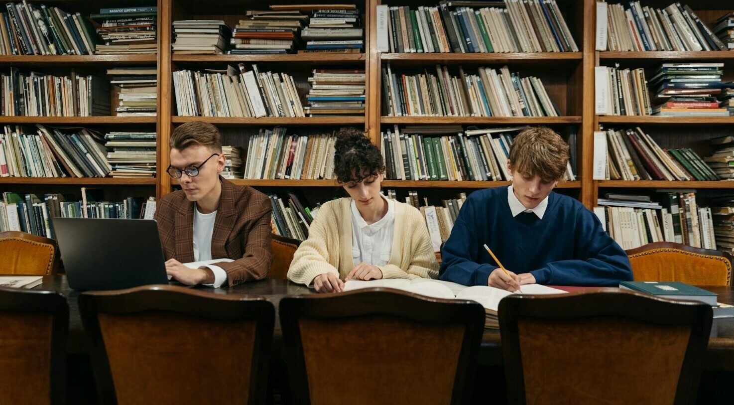 Three students studying at a library table, books behind them, one using a laptop.