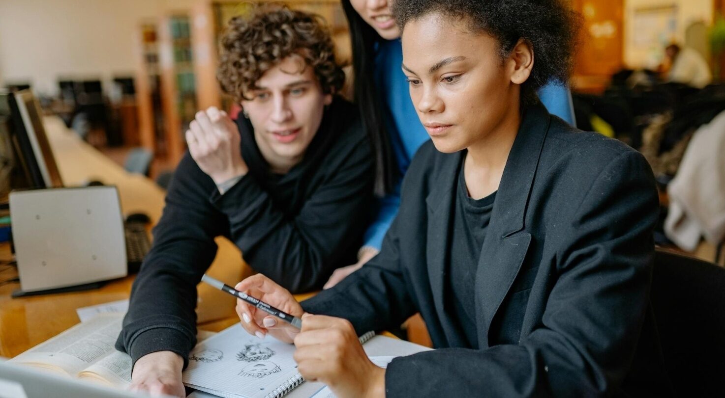 Three young people working together, looking at a laptop, with notes and sketches on the table.