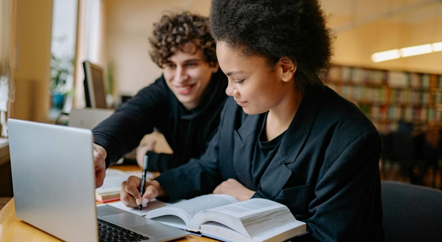 A boy and a girl studying together. The girl writes notes from a book, and the boy points at a laptop.