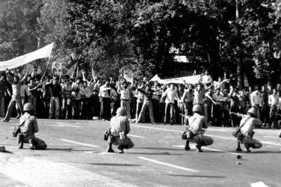 A large crowd of people protest while soldiers kneel with rifles in the street.