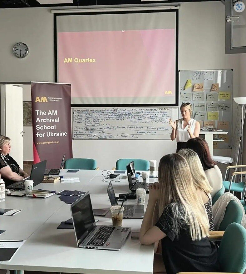 A woman presents with a projector screen displaying 'AM Quartex'. Attendees are seated at a table with laptops.