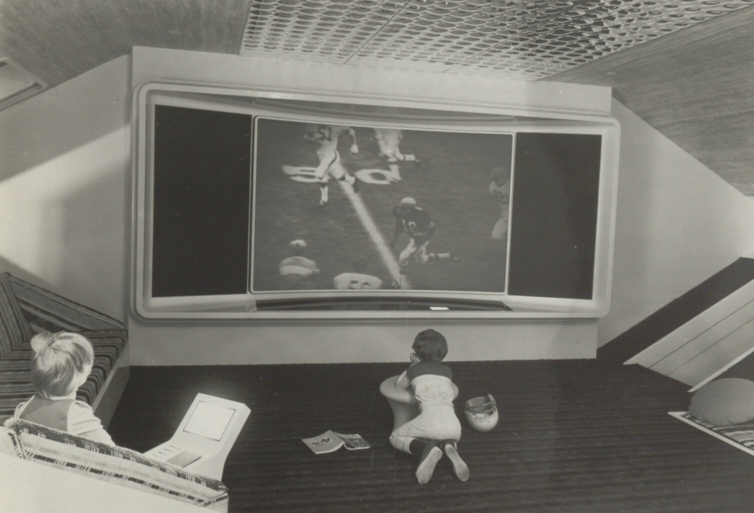 A child sitting on the floor watching a large TV screen while an adult sits nearby using a laptop