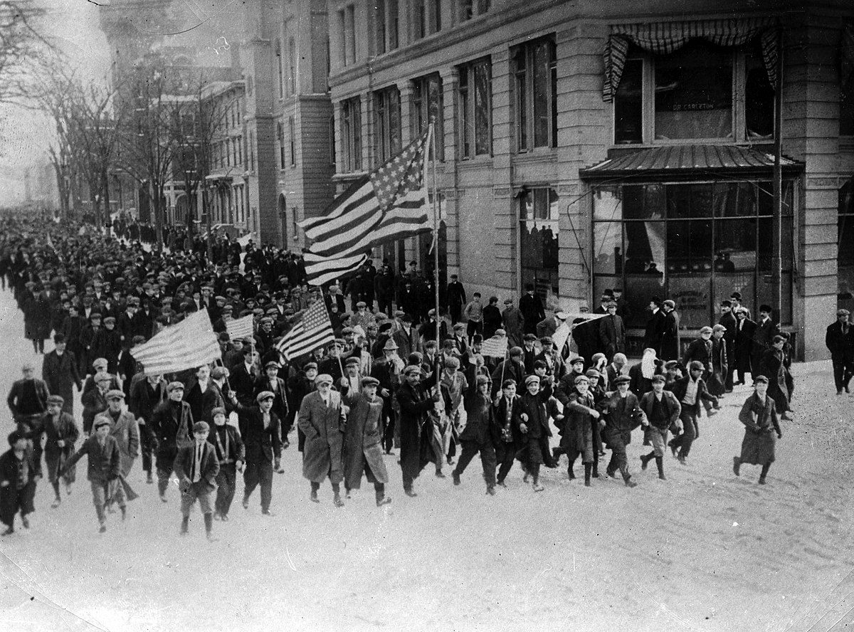 A large group of people marching in a parade, holding flags. Buildings line the street