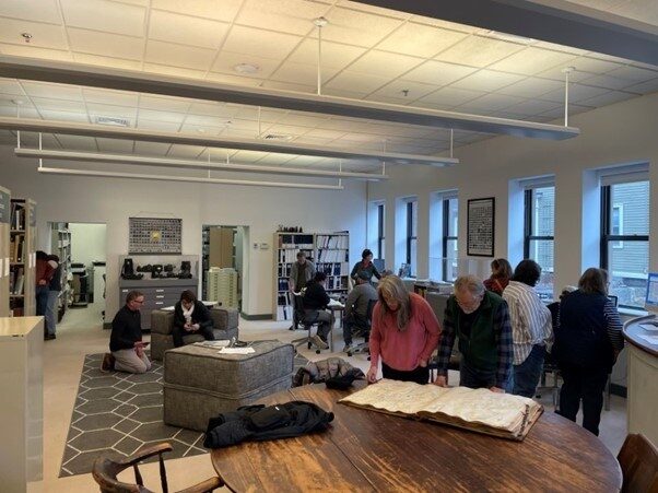 A group of people in a spacious room with bookshelves, sitting and looking at materials on a table.