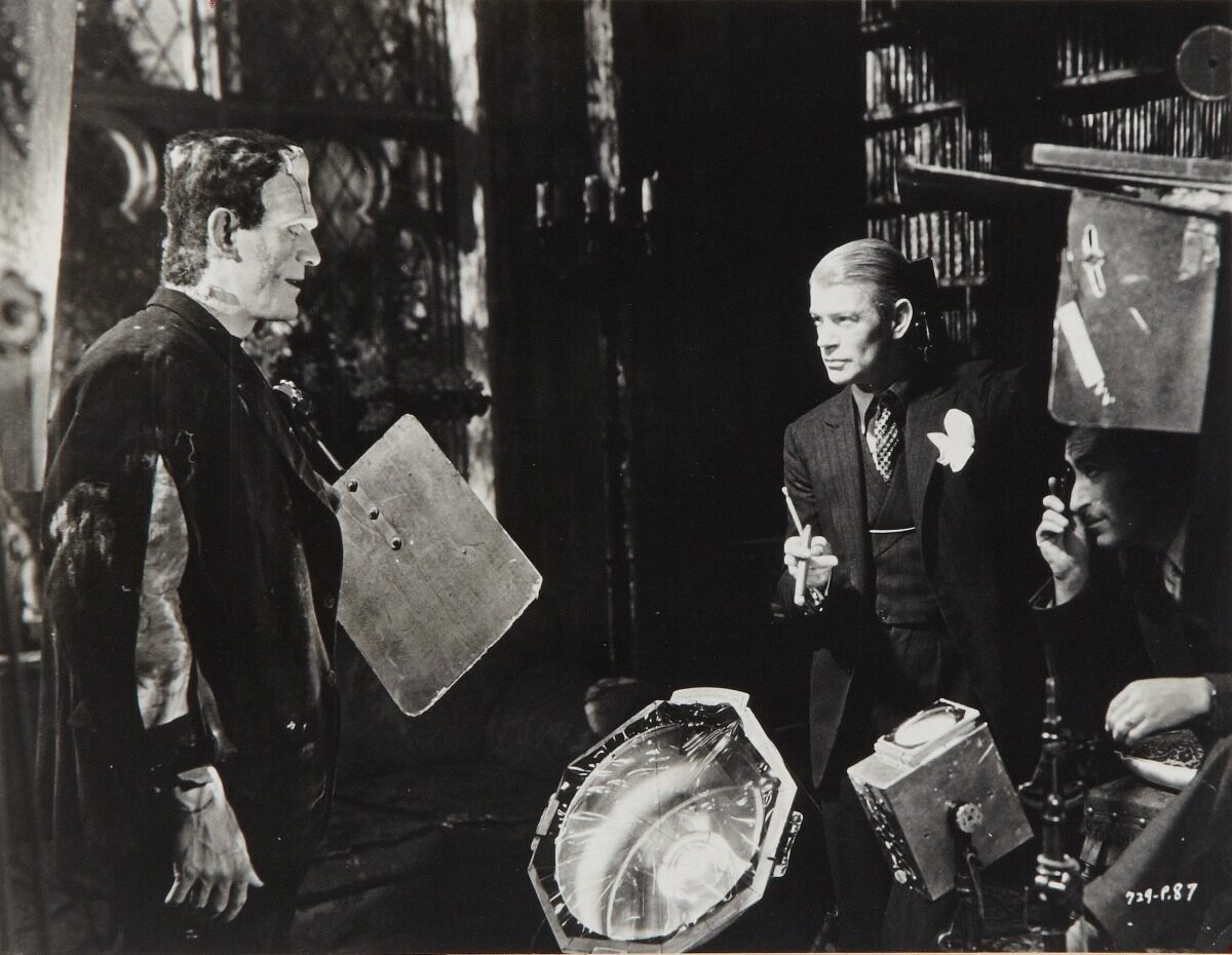 A scene from a classic film, featuring a monster and a man in a suit talking, with lighting equipment visible.