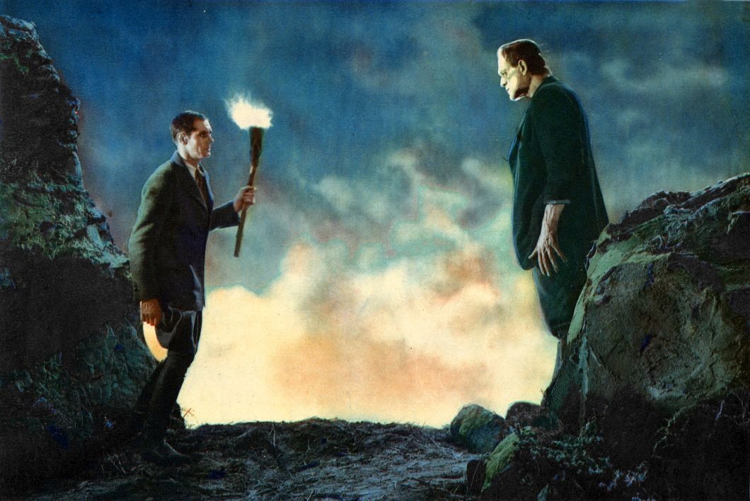 A man holding a torch stands on a hill looking up at a large figure in a suit.