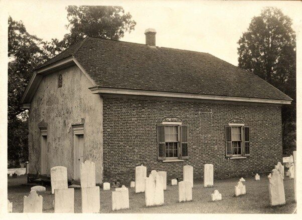 A small brick building with two windows, surrounded by tombstones in a graveyard.