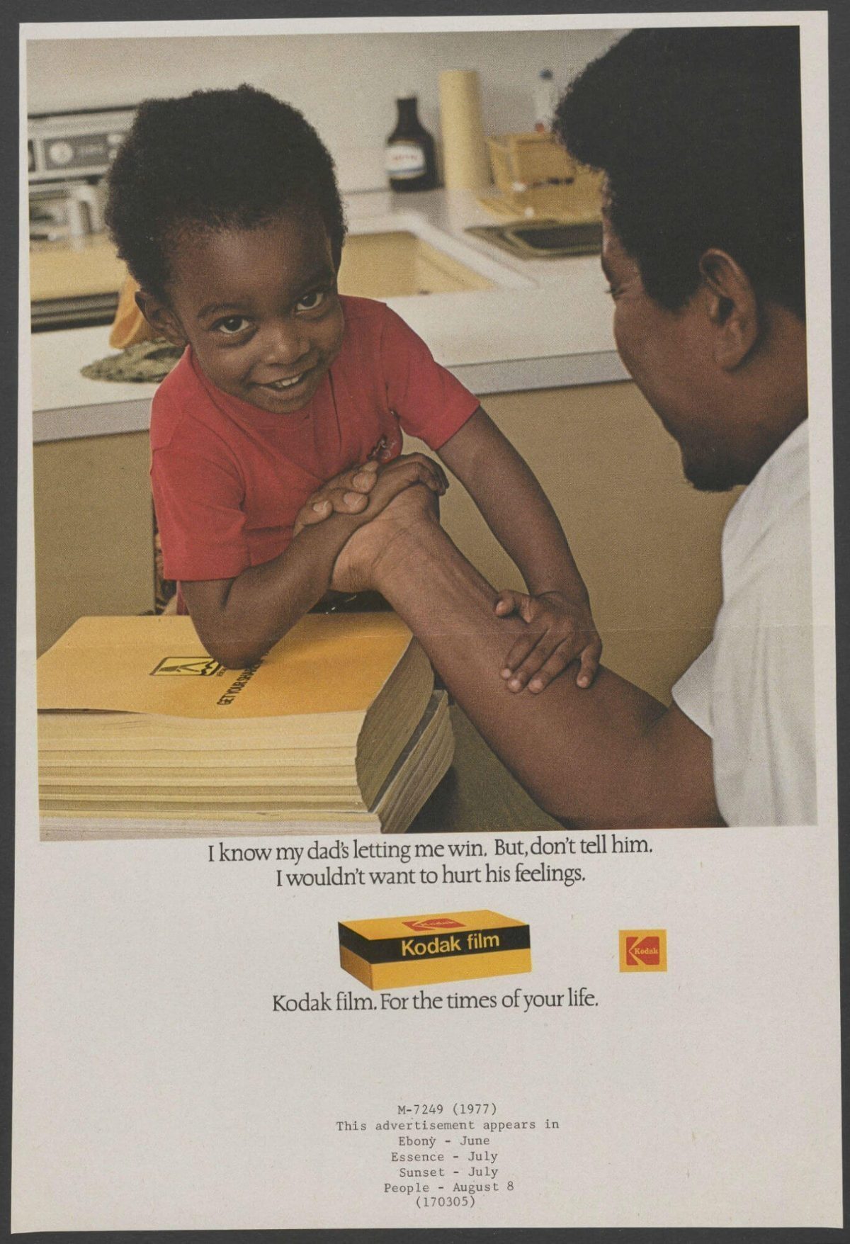 A young boy in a red shirt playfully arm wrestling with an adult man, surrounded by Kodak film products.