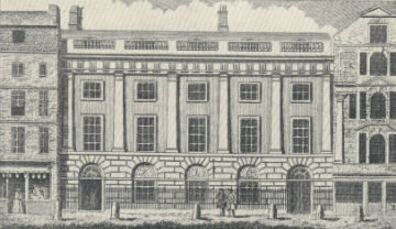 A historic building with columns and large windows, surrounded by older structures.