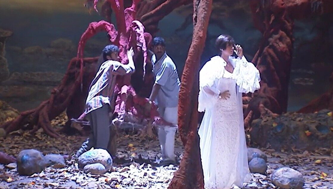 Two actors dressed in gray and white perform on stage beside a red tree, with a third actor in a long white dress