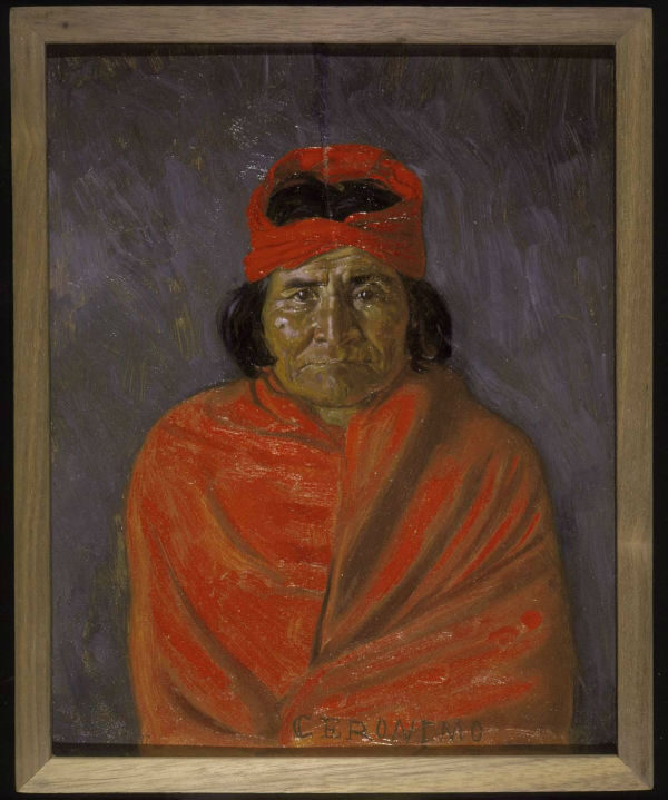A man wearing a red hat and orange blanket, with long black hair and serious expression.