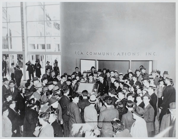 A large crowd of people gathered around a display at RCA Communications, dressed in 1930s fashion.