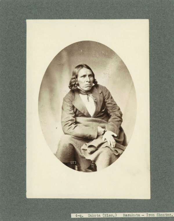 A seated man with long hair, dressed in nineteenth-century clothing, holding a blanket.