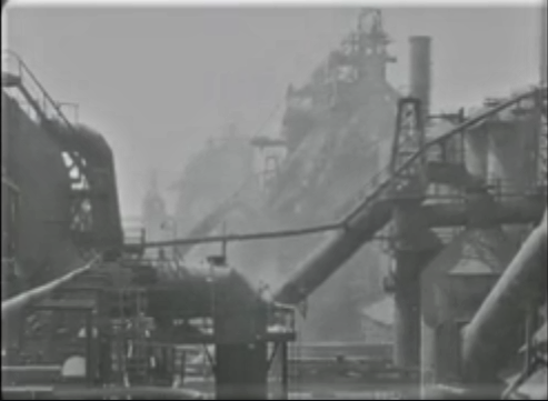 An industrial scene with large pipes and smokestacks, shrouded in fog.