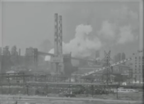A black and white photo of a large industrial complex emitting smoke, with tall chimneys and power lines.