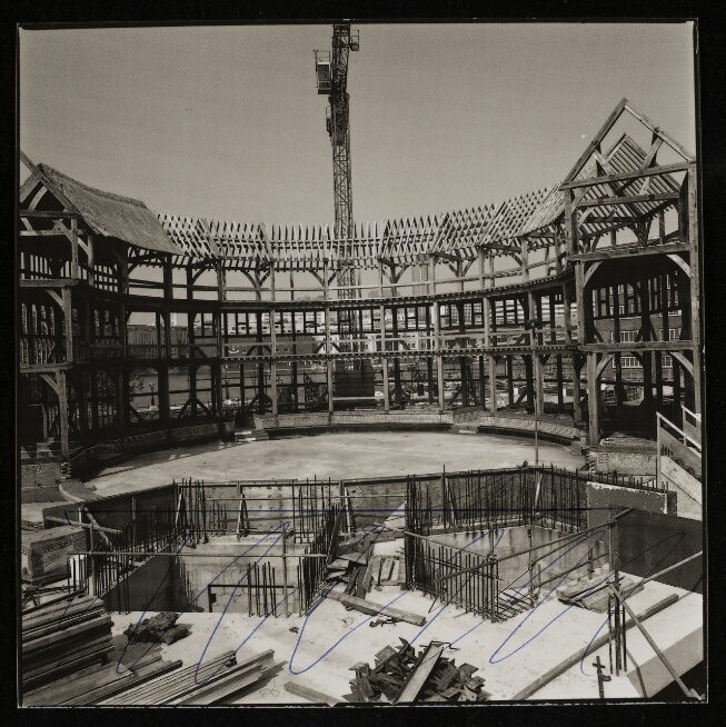 A black and white photo of a construction site with a circular wooden structure, metal reinforcements, and a crane.