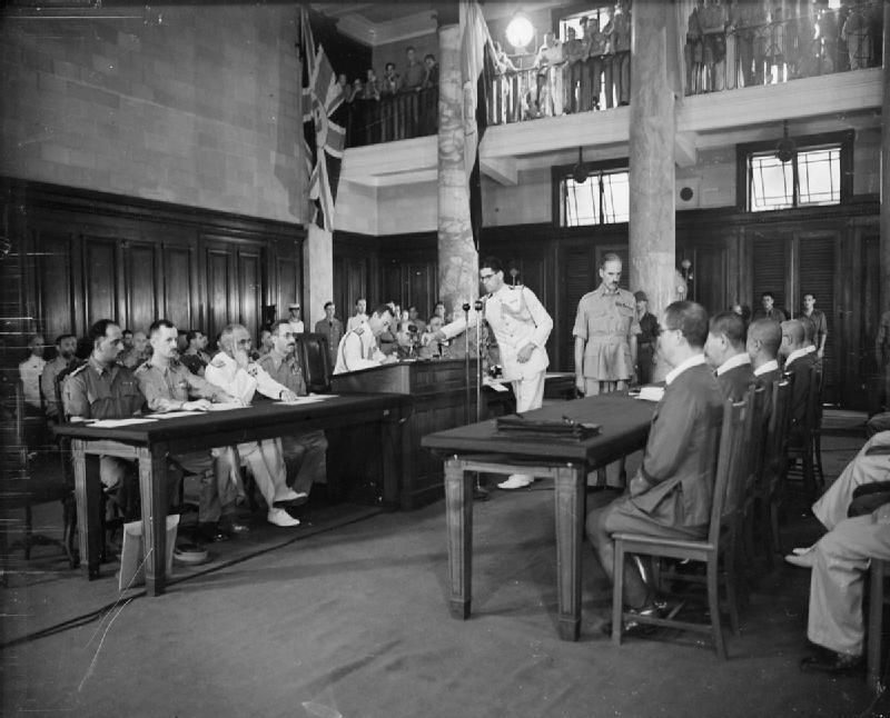 A historic courtroom scene with military officials and judges during a trial. Flags hang overhead.