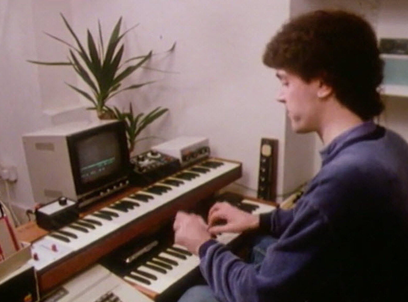 A young man plays a keyboard in front of a small monitor and a potted plant on a desk