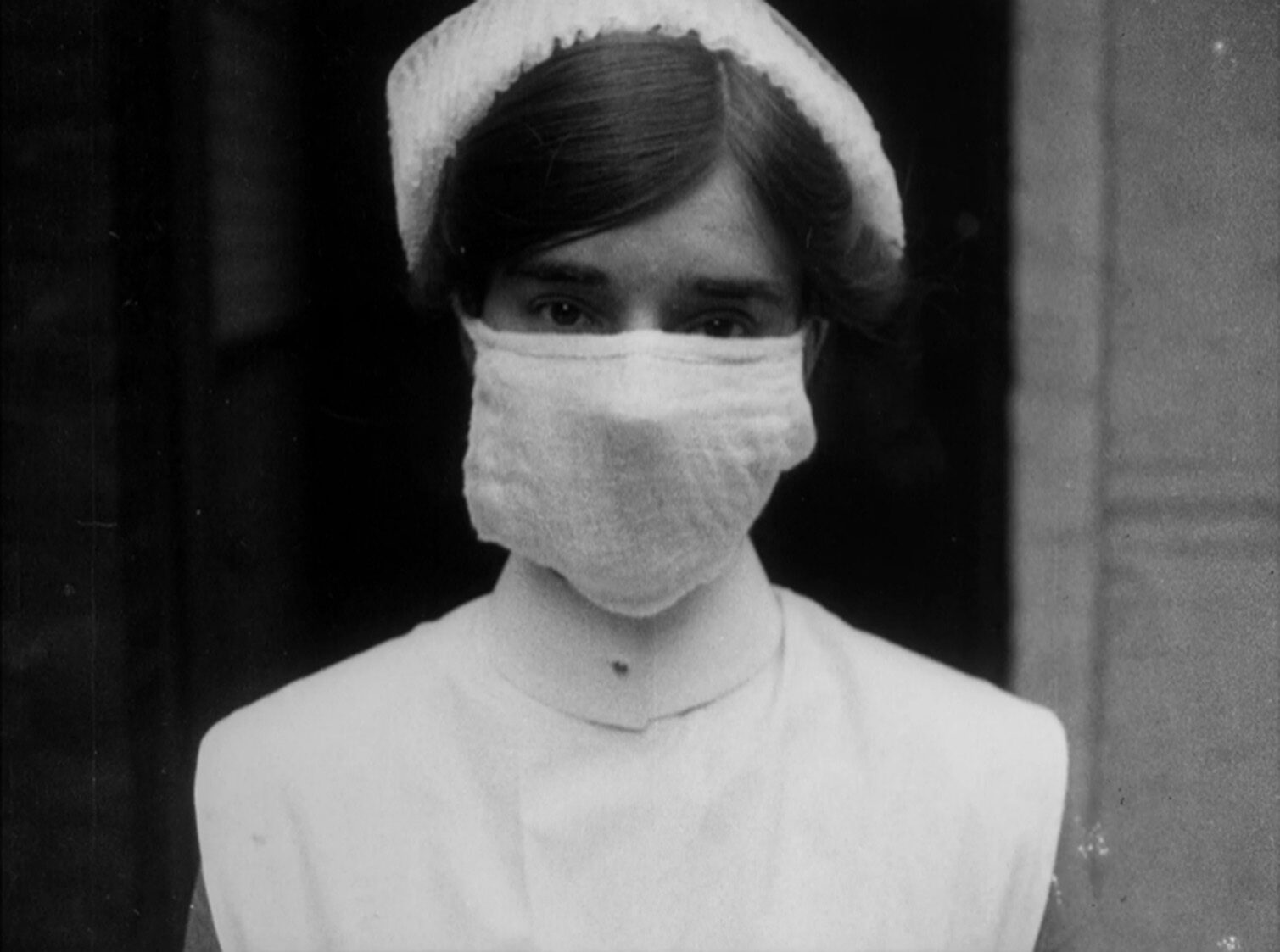A nurse wearing a fabric mask and a white cap, looking directly at the camera.