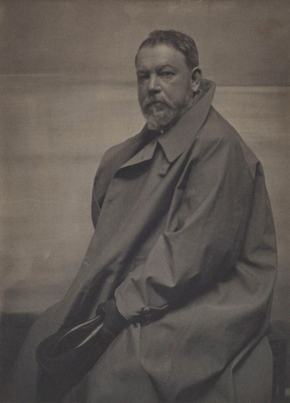 A man with a beard wearing a large trench coat, holding a black hat.