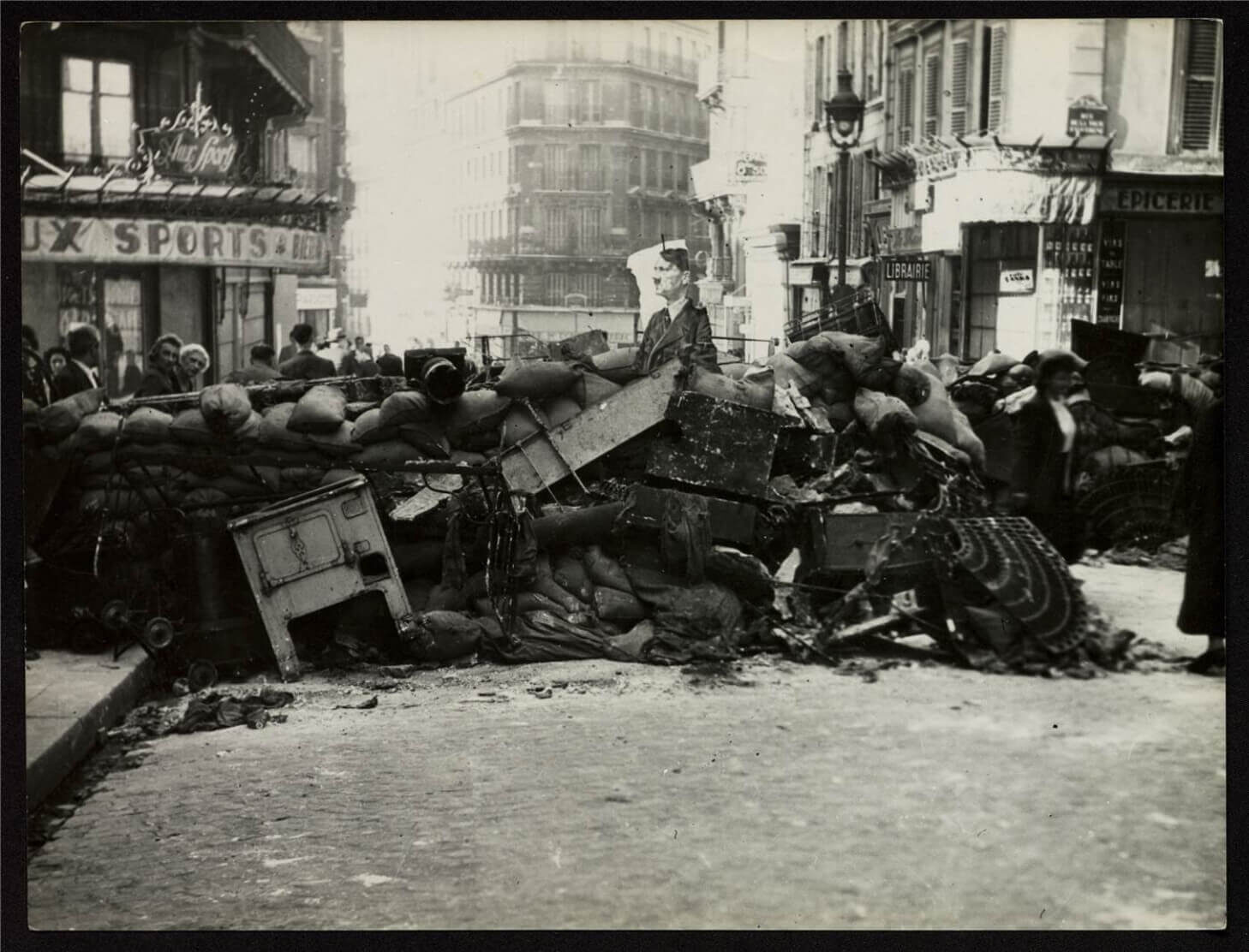 A historic street scene showing people gathering around barricades made of furniture and debris
