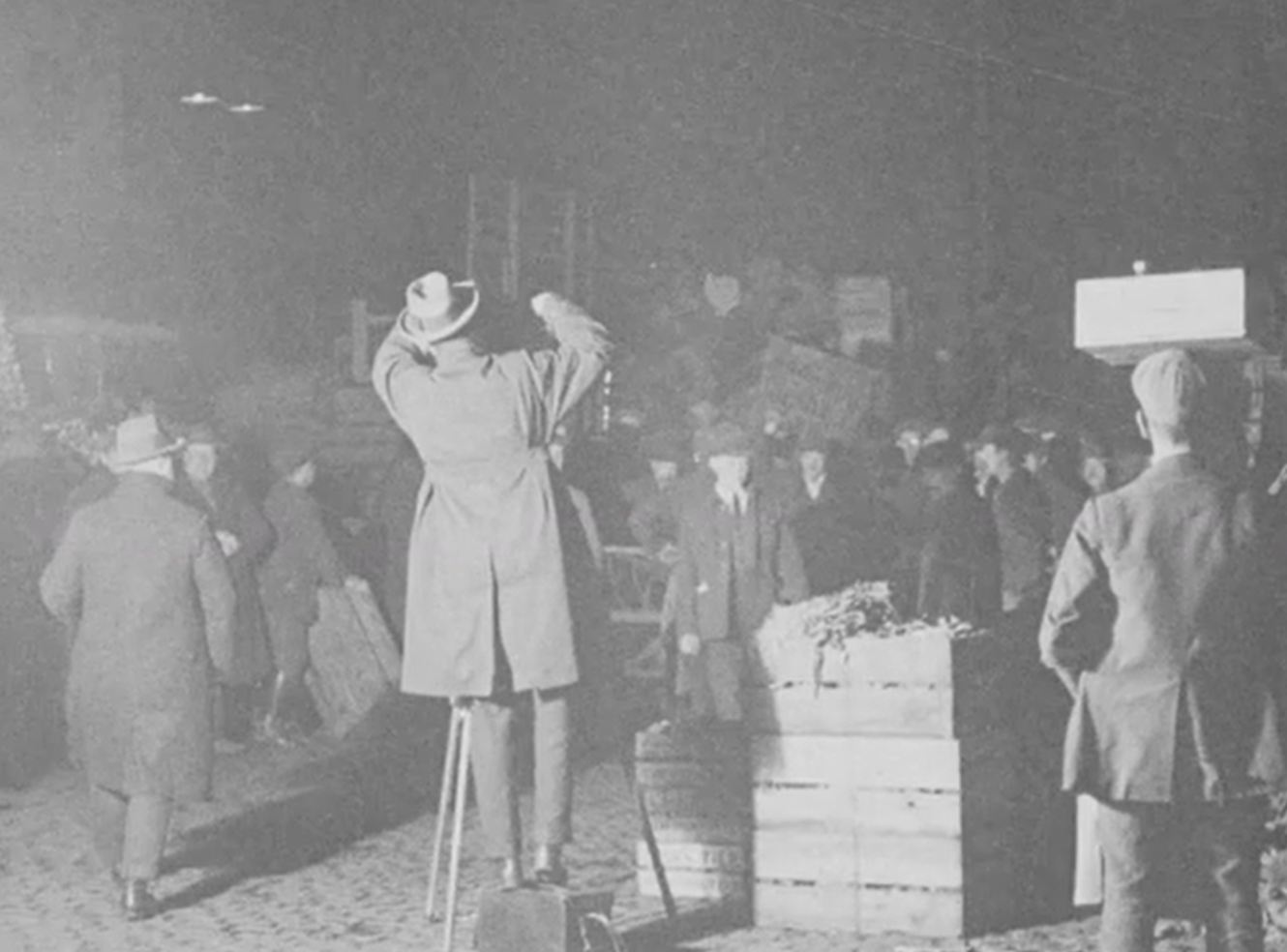 A man in a suit and hat stands on a crate, addressing a crowd at night