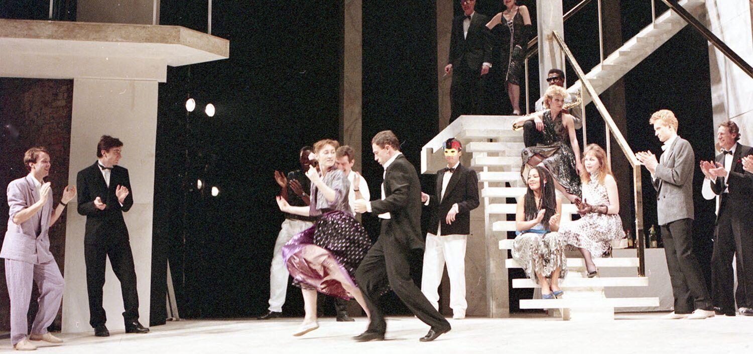 A lively stage scene with dancers and audience clapping, set on a grand staircase