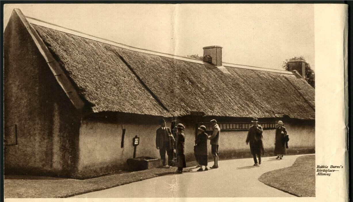 A thatched-roof house with six people standing outside, near a sign that mentions Robbie Burns.