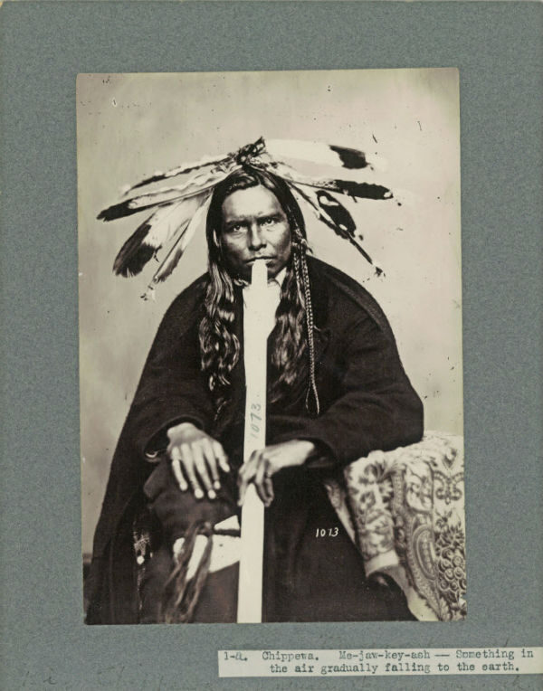 A man wearing a feather headdress plays a long flute, seated with hands on knees.
