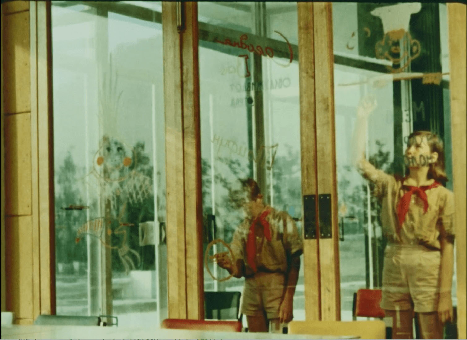 Two children in scouts uniforms cleaning glass doors, with green plants visible outside.