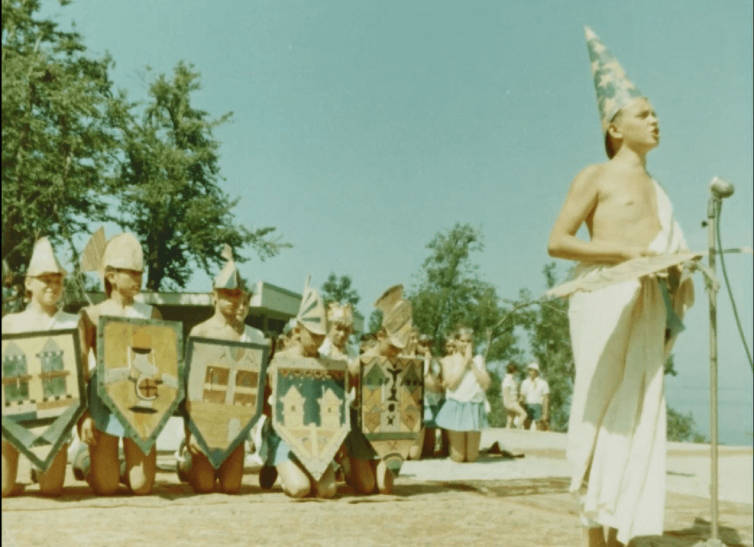 A young boy in a toga stands by a microphone, with six others kneeling behind, all in colourful costumes.