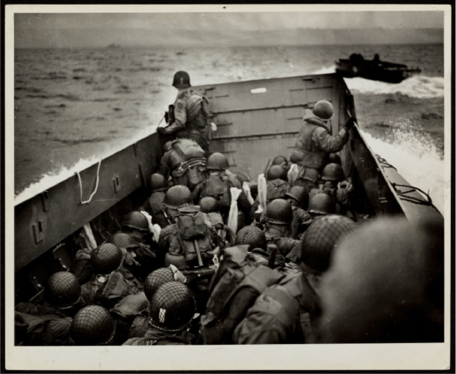 Soldiers in helmets and military gear sit and stand inside a landing craft at sea with another craft visible in the background under an overcast sky